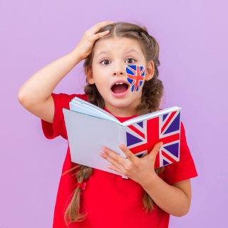 Kleines Mädchen mit Zöpfchen guckt erstaunt und fasst sich mit der Hand an den Kopf. Sie hält ein Buch mit englischer Flagge in der Hand, auf der Wange ist sie bemalt. Symbolbild für denglische Wörter und deren ganz andere Bedeutung auf Englisch.