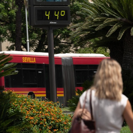 Eine Frau läuft neben einem Bus vorbei auf dem das Wort Sevilla steht. Über ihrem Kopf zeigt ein Thermometer die Temperatur von 44 Grad Celsius an.