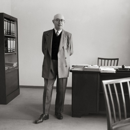 Die Aufnahme von 1958 zeigt den Philosophen Theodor W. Adorno mit hinter dem Rücken verschränkten Händen neben seinem Schreibtisch stehend. 