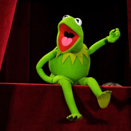Die Puppe "Kermit der Frosch" sitzt auf der Bühne mit roten Vorhängen an der Seite.