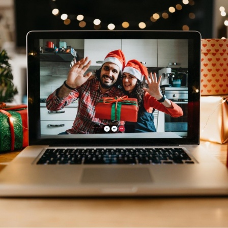 Ein junges Paar mit Weihnachtsmützen auf dem Kopf und Geschenk in der Hand winkt in die Kamera eines Laptop-Bildschirms.