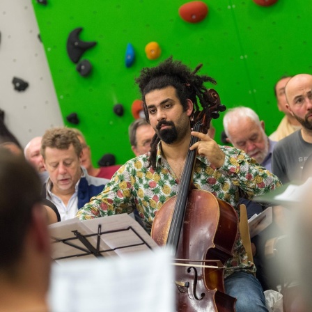 Akram spielt sein Cello, umringt von weißen Sängern, vor der Wand einer Kletterhalle.