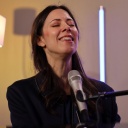 Eine Sängering mit langen dunklen Haaren sitzt lächelnd mit geschlossenen Augen an einem Mikrofon.