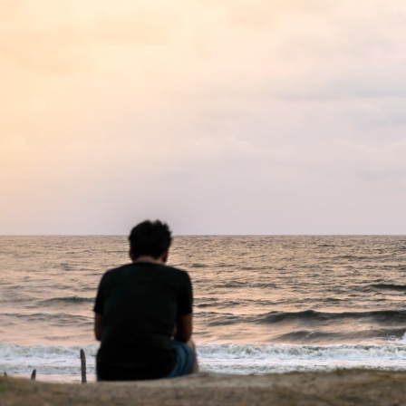 Eine Person sitzt leicht zusammengesunken alleine am Meer.