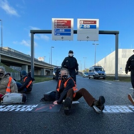 Klimaschutz-Aktivisten der Initiative "Aufstand der letzten Generation" blockieren eine Zufahrt zum Hauptstadt-Flughafen BER. Auf dem Boden liegen Banner mit der Aufschrift «Essen retten, Leben retten».