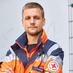 Ein Mann Mitte 40 mit kurzen Haaren und Kinnbart, Tobias Schlegl, steht in der Jacke eines Notfallsanitäters vor einer weißen Wand und blickt in die Kamera.