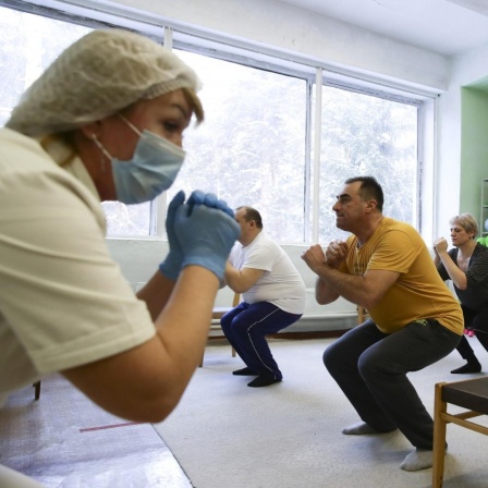 In einem Raum machen mehrere Menschen eine Kniebeuge. Vor ihnen steht eine Krankenschwester mit einem Mundnasenschutz.