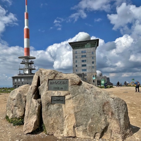 Eine weißrote Antenne, davor ein Felsen, auf dem eine Plakette mit der Inschrift "Brocken" steht, dahinter ein hohes Gebäude