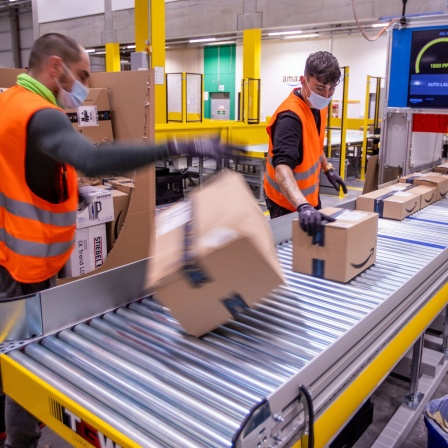 Zwei Männer legen Pakete auf ein Band im Verteilzentrum des Online-Händlers Amazon in Mecklenburg-Vorpommern: Der Anteil von Amazon am Online-Markt liegt in Deutschland bei 55 Prozent, in den USA bei über 70. Missbraucht der Konzern seine Marktmacht zum Nachteil des Wettbewerbs und diktiert die Preise?