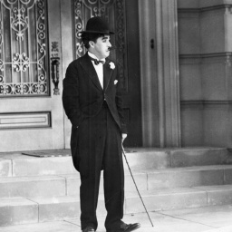 Charlie Chaplin steht vor einem Gebäude und hat einen Gehstock in der Hand. 