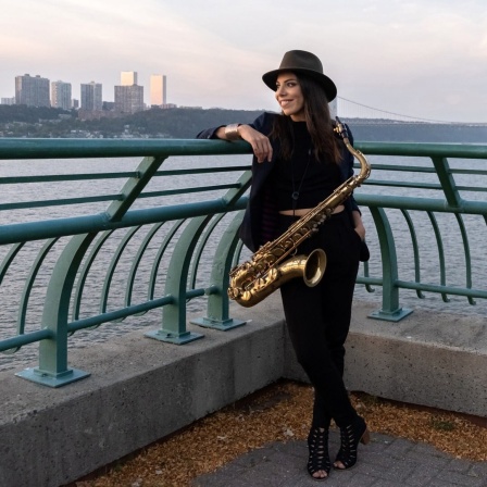 Die Chilenin Melissa Aldana steht mit ihrem Saxofon an einem Geländer vor einem Gewässer, im Hintergrund eine Stadt Skyline.