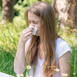 Eine junge Frau sitz im Gras und schnäuzt sich die Nase in ein Taschentuch.