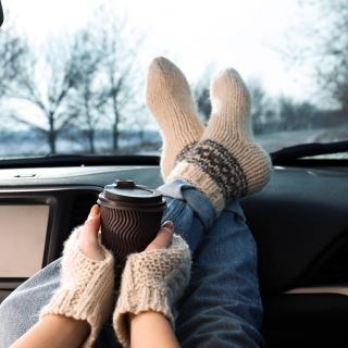 Junge Frau in warmen Socken hat ihre Beine aufs Armaturenbrett eines Autos gelegt und hält einen Becher Kaffee oder Tee: Tee und Kaffee sorgen für Harndrang und kalte Füße. Das liegt am Koffein.
