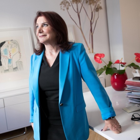 Eine dunkelhaarige Frau in blauem Blazer und schwarzem T-Shirt lehnt sich an einen Schreibtisch, auf dem unter anderem mehrere Akten liegen. Sie blickt zur Seite. Die Frau ist die Rechtsanwältin Renate Maltry,