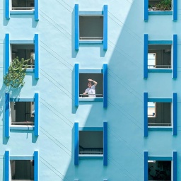 Ein Mann am Fenster eines Hauses mit blauer Fassade schützt seine Augen vor der Sonne.