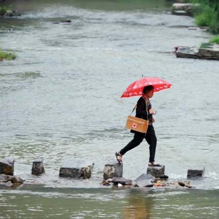 Eine Ärztin in China ist auf dem Weg in ein Nachbardorf: Zu sehen ist eine Frau, die über Schrittsteine durch einen Fluss geht, über der Schulter einen Medizinkoffer mit einem roten Kreuz darauf
