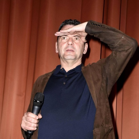 Christian Petzold steht vor einem Vorhang auf einer Bühne und schaut mit einer Handfläche an die Stirn gelegt in die Ferne.  