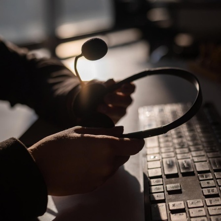 Dunkles Bild: Zwei Hände halten ein Headset über einer Tastatur.