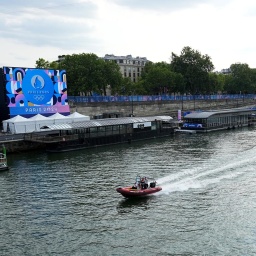 Ein Schlauchboot fährt vor den Olympischen Sommerspielen in Paris 2024 auf der Seine entlang. Am Ufer sind Aufbauten für Olympia zu erkennen.