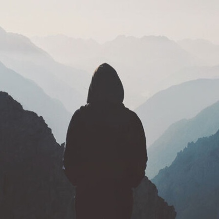 Ein Mann steht einsam auf einem Gipfel und blickt in ein Gebirgspanorama.