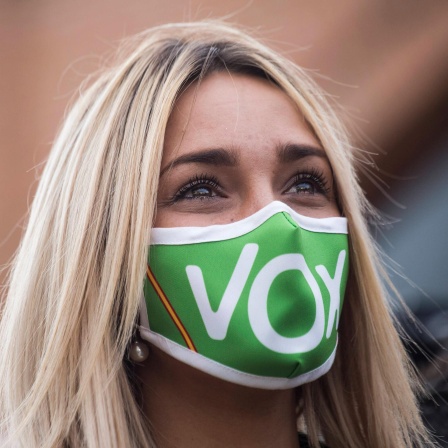 Eine junge blonde Frau trägt eine grüne Maske, auf der mit weißen Buchstaben Vox steht.