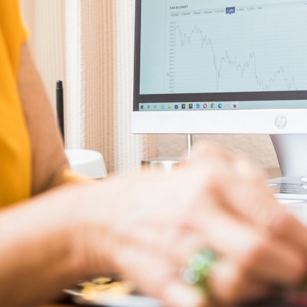 Die Hand einer Frau ist an einem Schreibtisch zu sehen, auf dem ein PC steht, der Aktien-Charts anzeigt.