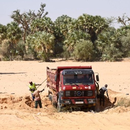 Ein Lastwagen auf sandigem Untergrund, Arbeiter schaufeln Sand in das Fahrzeug. Im Hintergrund sind Bäume zu sehen.