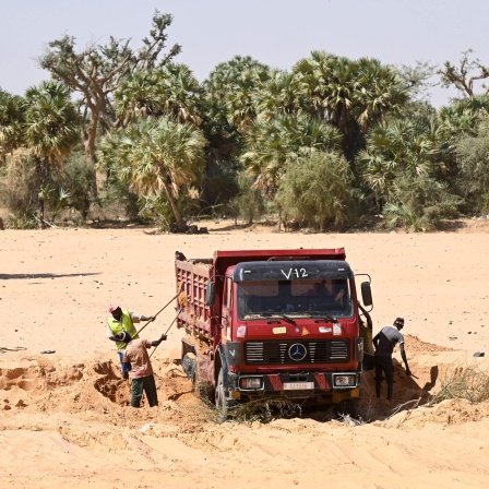Ein Lastwagen auf sandigem Untergrund, Arbeiter schaufeln Sand in das Fahrzeug. Im Hintergrund sind Bäume zu sehen.