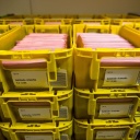 Zahlreiche Behälter mit Stimmzetteln der Sozialwahl stehen in einer Lagerhalle in gelben Kästen der Deutschen Post.