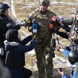 Ein Militär der ukrainischen Armee gibt ein Interview; er ist von Reporterinnen und Reportern umgehen. Alle stehen auf einem Feld. (20.02.2023)