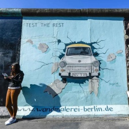 Eine junge Frau steht lesend vor der Berliner Mauer. Darauf ist eine Wandmalerei: ein Trabi, der die Mauer durchbricht.