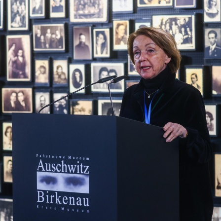 Eva Umlauf steht vor einer Wand mit Schwarz-weiß-Porträtfotos an einem Rednerpult mit der Aufschrift "Auschwitz Birkenau".