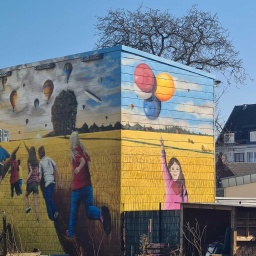 Mural in Köln-Ossendorf: Kinder laufen ausgelassen über ein gelbes Weizenfeld, über ihnen Ballons im blauen Himmel