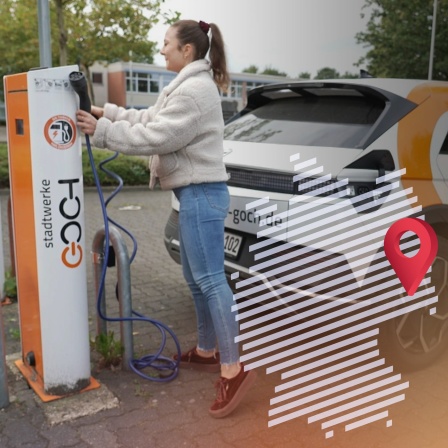 Eine junge Frau mit brauenm Haar, weißer Jacke und Jeans lädt das elektrische Dorfauto der Stadt Goch an einer Ladestation auf.