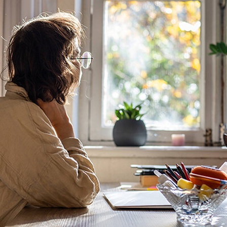 Foto einer jungen Frau, die nachdenklich am Schreibtisch sitzt und aus dem Fenster guckt