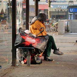 Eine wohnungslose Frau sitzt im Stadtteil Steglitz mit ihrer wenigen Habe an einer Bushaltestelle. Auf einer grossen Plastiktuete von ihr steht dabei "Home Discount". 