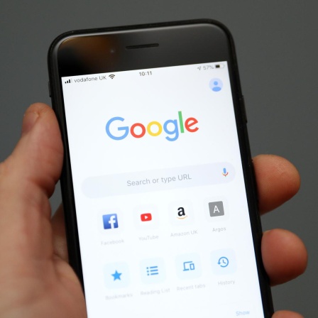 Eine Hand hält ein Mobiltelefon auf dessen Display die Oberfläche der Suchmaschine Google zu sehen ist. 