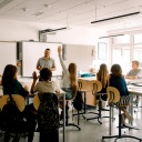 Schüler sitzen in einem Klassenraum. Vorn steht ein Mann mit dunklen, kurzen Haaren, der die Gruppe anlacht.