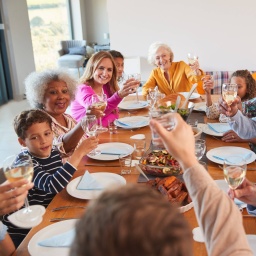 Bunte erweiterte Familie sitzt um gedeckten Tisch und erhebt Gläser mit Wasser und Weißwein. Freudige Gesichter. Blick zur Person an Stirnkante, unscharf mittig im Vordergrund.
