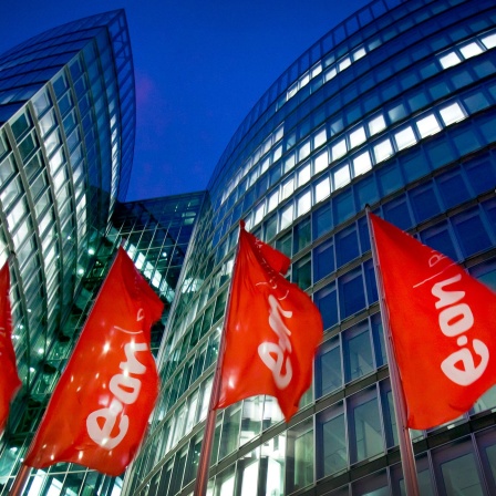 Die Zentrale von Eon-Ruhrgas in Essen, rote Fahnen mit Firmenlogo wehen vor einem hohen Gebäude mit beleuchteten Fenster vor dunkelblaumen Himmel