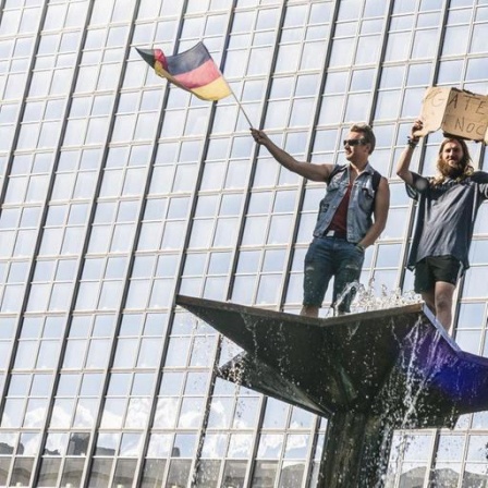 Zwei Demonstranten bei einer "Hygienedemo"protestieren auf dem Brunnen der Völkerfreundschaft auf dem Alexanderplatz in Berlin. Einer hält ein Schild: "Gate's noch?", ein anderer schwingt die Deutschlandfahne, 9. Mai 2020.