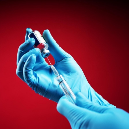 Zwei Hände in blauen Handschuhen ziehen vor rotem Hintergrund eine Spritze auf.