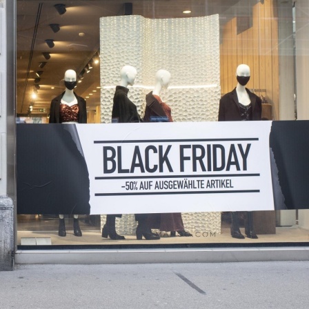 Eine Frau steht mit Maske vor einem Schaufenster, das für den "Black Friday" geschmückt ist. 