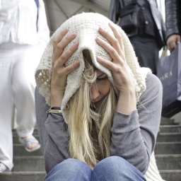 Junge Frau sitzt verloren mit einem Schal über dem Kopf alleine auf einer Treppe, während Menschen an ihr vorbeigehen