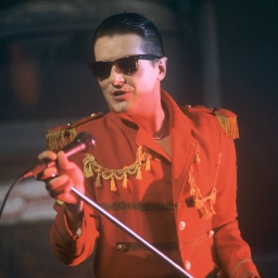 Sänger Falco singt in roter Jacke, mit Sonnenbrille und zurückgegelten Haaren in ein Mikrofon, dass er mitsamt Ständer hohgehoben hat.