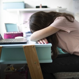 Ein Mädchen sitzt am Schreibtisch, vor sich einen Laptop, und hat den Kopf auf die Arme gelegt.