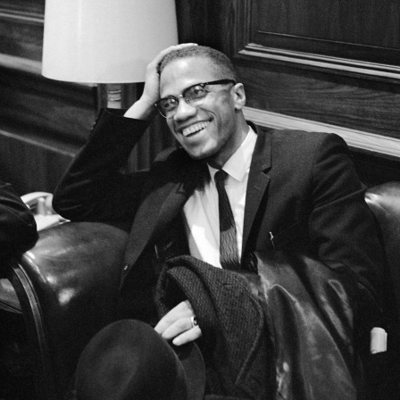 Der US-Bürgerrechtler Malcolm X im Anzug, er sitzt lächelnd auf einem Sessel und wartet auf die Pressekonferenz von Martin Luther King im Jahr 1964.