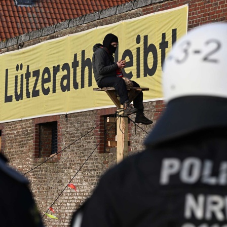 Ein Demonstrant sitzt bei der Räumung des Dorfes Lützerath auf einer Holzkonstruktion vor einem Plakat mit dem Slogan "Lützerath bleibt!" Im Vordergrund sind polizisten zu sehen. (Quelle: dpa)