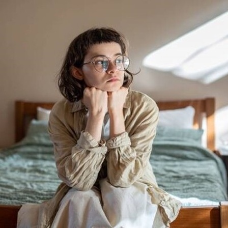 Eine Frau sitzt auf einem Bett und guckt deprimiert aus dem Fenster