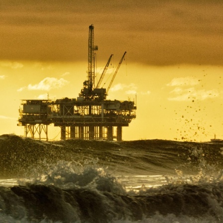 Gelblich gefärbte Meereslandschaft bei Sonnenauf- oder -untergang: Im Vordergrund hohe, schäumende Wellen, dahinter ragt eine große Offshore-Bohrplattform mit Kränen und Stützpfeilern aus dem Wasser, darüber ein wolkiger Himmel.